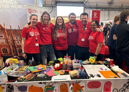 Lab members in red Queen's University Belfast t-shirts behind a stand covered in craft materials and children's drawings of viruses